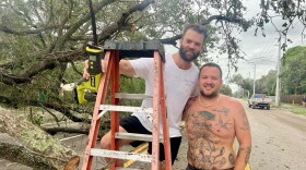 One man stands on a latter holding a chainsaw. Another man stands next to him. They're under a large oak tree that fell during Hurricane Milton.