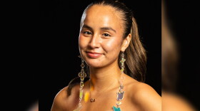 A woman poses against a black background. She is wearing a pink strapless dress and long beaded earrings. 