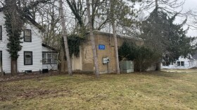 On a patchy, muddy lawn, a small brick building sits surrounded by trees and covered in moss. The building has no windows, just a small door and a blue plaque with the numbers 1509
