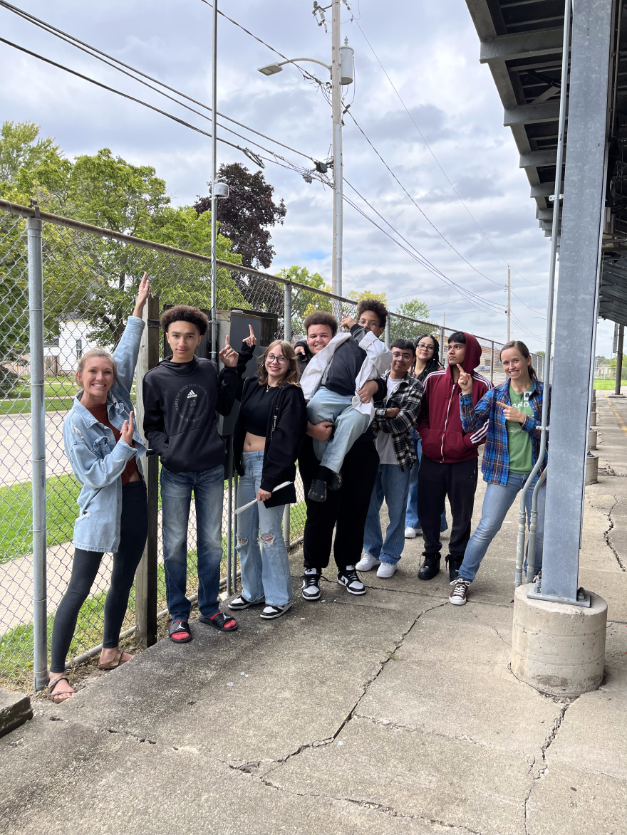 a group of diverse high schoolers smiles outside and points up to a small white device