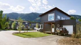 Grey tiny houses surrounded by green mountains and trees, with a Jeep parked by a white garage door.