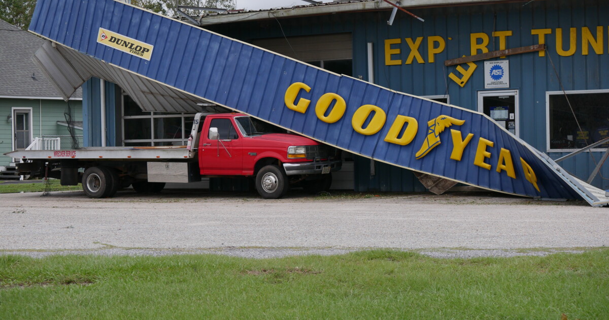 A collapsed blue Goodyear storefront facade rests across a red flatbed truck outside a damaged tire shop.