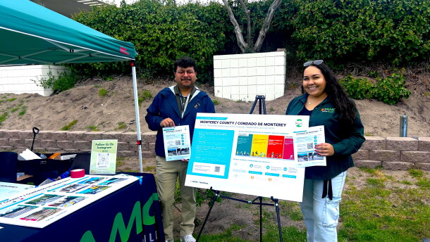 Two people standing holding flyers with a poster board on a stand between them. They are standing outside, a booth with a pop-up tent is to their left.
