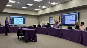 A man stands in front of a group assembled around a board room, presenting on a large screen. 