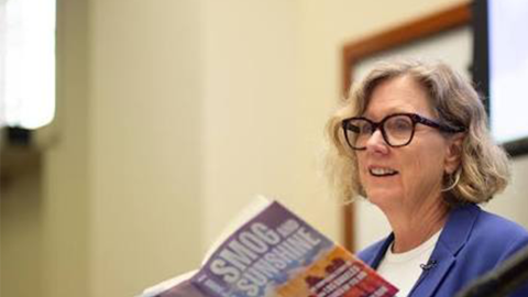 A woman with short blonde hair and black glasses wears a blue blazer and reads a book titled "Smog and Sunshine"