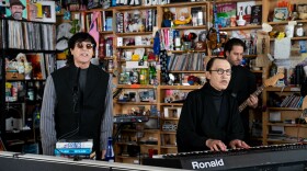  A man wearing sunglasses sings into a microphone while standing behind an office desk as another man wearing spectacles plays a keyboard and a third man in the background plays bass guitar.