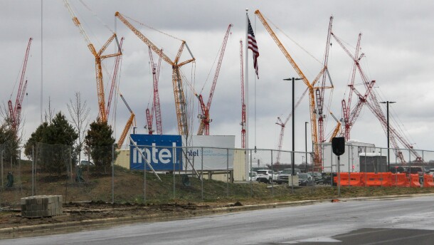 Large, metal cranes rise above an entrance to a construction site. A sign, partially obscured by a chain-link fence, reads, "Intel." 