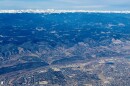 Dry mountains and hills are pictured next to sprawling cities as seen from an aerial photograph. A small ribbon of snow capped mountains are in the background. 