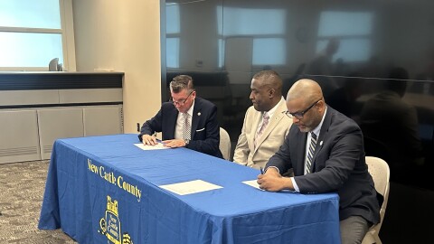 Three men sit at a table and sign papers. The table cloth has the New Castle County emblem. 