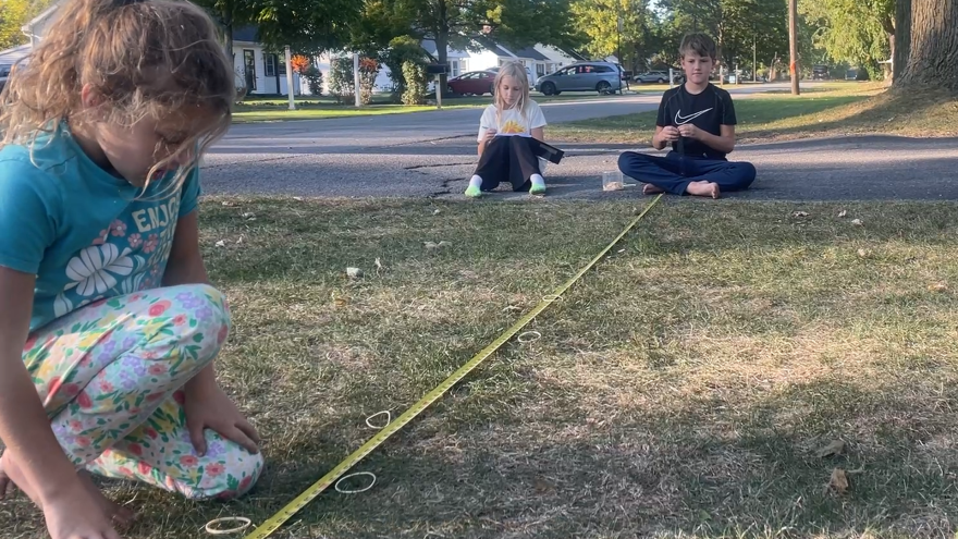 In the foreground is a young girl kneeling in the grass, looking at the flung rubber bands and measuring their distances with a tape measurer. In the background are a young girl and a young boy sitting on the blacktop.