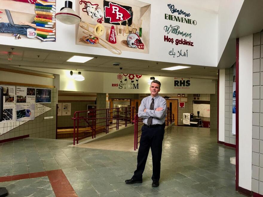 A man stands in a high school atrium below a banner with a red arrowhead with two r's inside of it