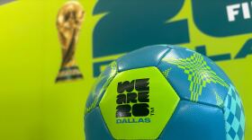 A blue patterned soccer ball printed with "We Are 26 Dallas" sits in front of a lime green backdrop to promote the 2026 FIFA World Cup. 