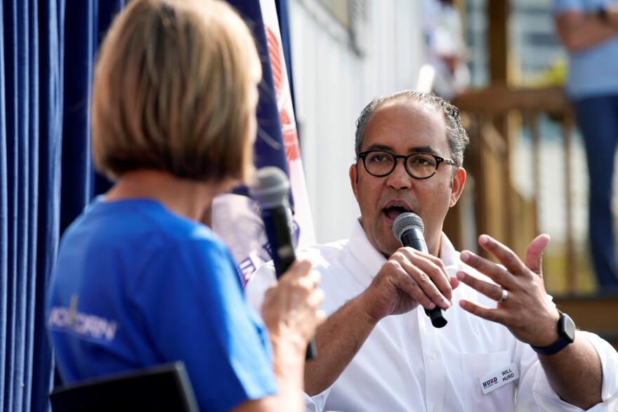 A man holds a microphone as he speaks to a woman who's back is to the camera.