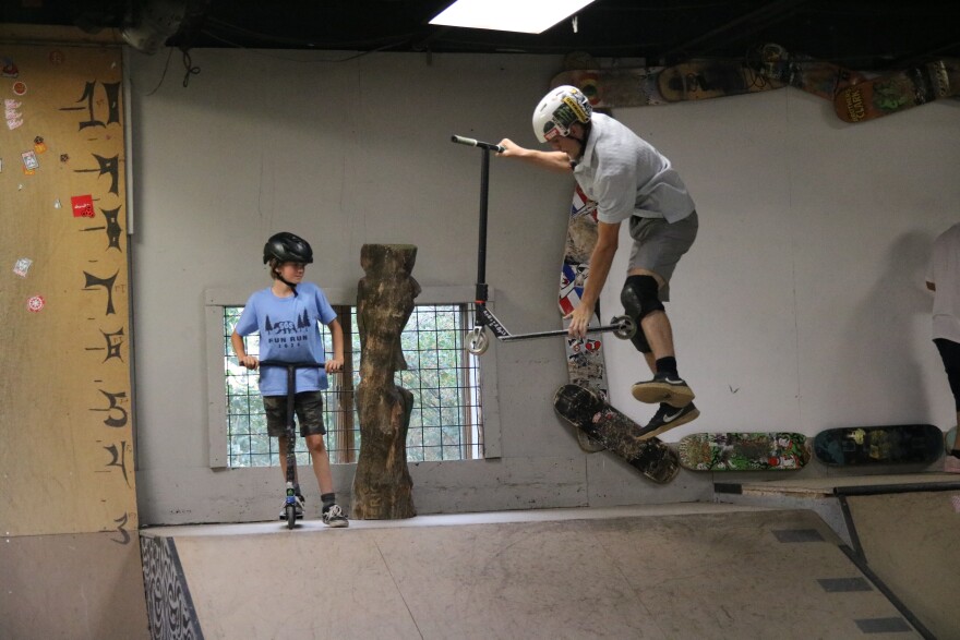 A guy in a white helmet, grey-blue shirt and shorts grabs his handlebars of his scooter with one hand and the stand with the other while hanging midair. A boy in a blue shirt and shorts with a black helmet watches him from the top of a quarter pipe.