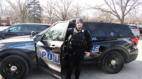 A police officer stands in front of the driver's side of his squad car. The door is partially open and he is behind it with his hand on the door. 