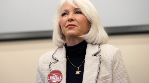 Tin Peters with white hair, wears a white jacket over a black shirt. She has a red button with her own name on it.