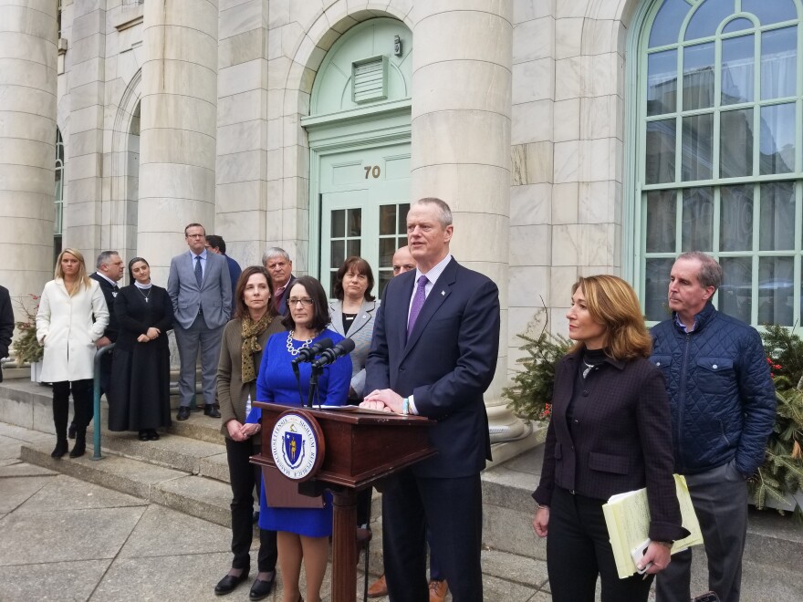 Massachusetts Governor Charlie Baker stands at a podium in front of a marble building with a row of people standing on either side of him