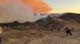 A string of 8 firefighters appear on a dirt mountain as fires blaze on green rolling hills behind them. 