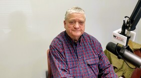 A man in a plaid, long-sleaved button down shirt sits in a radio studio, smiling at the camera.