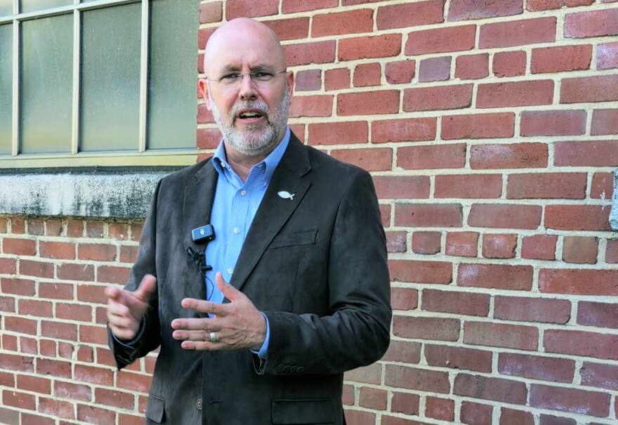 A man in a suit stands in front of a brick wall and talks with the media.