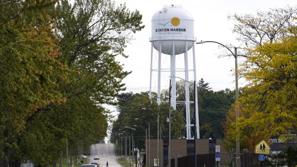 A lone resident of Benton Harbor walks across a street near the city's white water tower, with the city's name plastered on the top of the tank. A few cars can be seen driving towards the viewer, with the orange tinged leaves of the trees surrounding the street denoting the Autumn months.