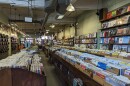 A record store has long aisles filled with albums, with additional albums on wall shelves and 7-inch singles hanging from pegs.