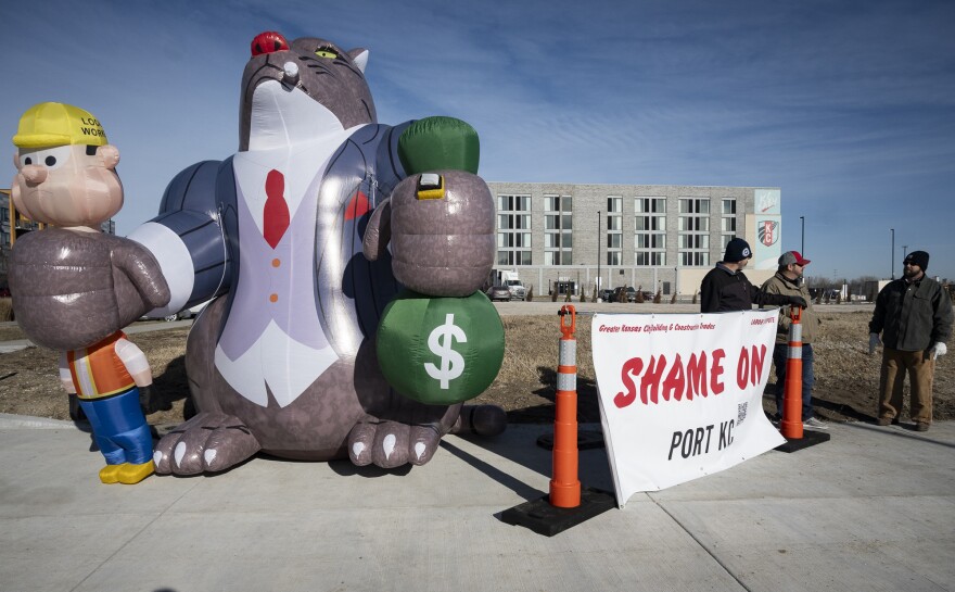 Three men can be seen standing outdoors on a corner of an intersection. They are standing behind a banner that reads "Shame on Port KC." At left is a large inflatable cat that is is smoking a cigar wearing a suite. It  has its paws wrapped around an inflatable person who looks like a construction worker in its right paw and a bag of money in its left paw.