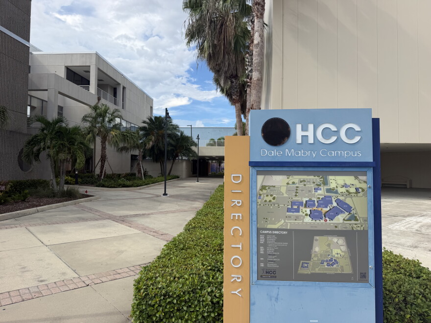 A blue and tan directory sign with a map of Hillsborough College's Dale Mabry campus stands in front of the entrance of grey classroom buildings and trees lining the walkway.