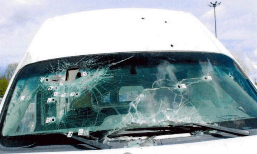 The front windshield of a white van, heavily cracked and pierced by multiple bullet holes with numbered police evidence markers placed next to the impact points.