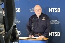 This is a photo of NTSB Board Member Tom Chapman speaking at a lectern in front of a single microphone during a press conference in Cheektowaga. Chapman is older man with a beard, no hair and glasses. He's wearing a navy blue button-up emblazoned with the NTSB's logo on it. A television camera can be seen in the left side of the image. 