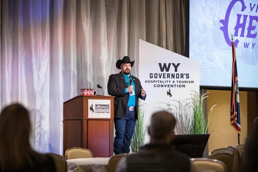 A man wearing a cowboy hat and a black blazer speaks into a microphone as he stands next to a podium and a sign that reads “WY Governor’s Hospitality & Tourism Convention.”