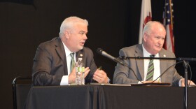 Two men in suits sit at a table on a stage with a microphone between them.