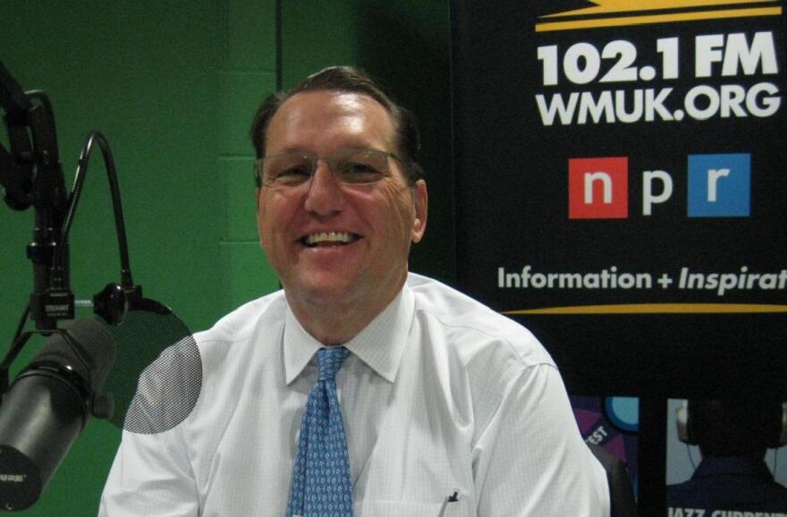 A head and shoulders photo of a man with dark hair, glasses and a tie, who is smiling and sitting behind a microphone. A colorful banner for WMUK is in the background. 