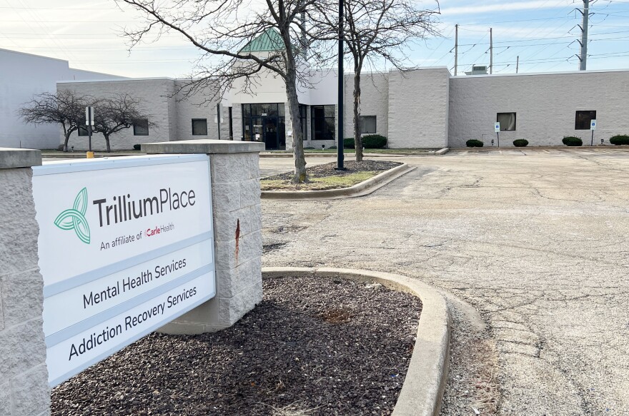 An entry sign identifying Trillium Place sits on a median in a driveway in front of a large medical office building with a green roof, partially obscured by trees and a light post on another median.  