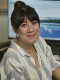 A person sits, smiling and looking at the camera. They are sitting in front of a computer screen.