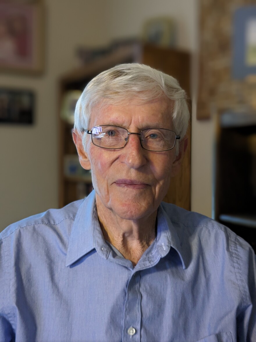 A photo of John Ikerd. He is seated inside and is wearing a light blue shirt and glasses.