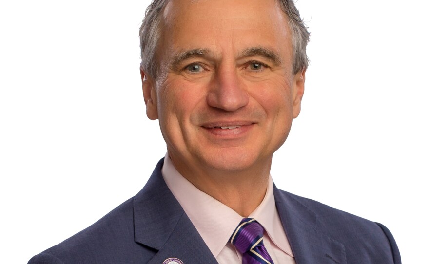 A man with short grey hair is wearing a navy blazer, pink button-down shirt and purple striped tie.