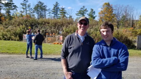 Two men stand in front of a man and a woman in a field. The woman is holding up a fun at a row of targets.