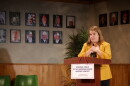 A woman wearing a yellow coat standing in front of a podium with pictures of military servicemen in the background.