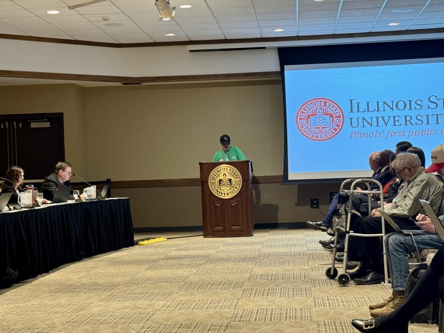 A person speaks at a podium with the Illinois State University seal, while people sit and listen. The university’s logo and motto are projected on a screen behind them in a conference room setting.