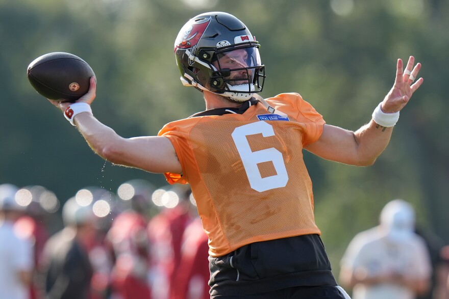 A white man wearing an orange football jersey with the number 6 on it in white font and a dark gray football helmet with the red, white, and black buccaneers flag logo. He has both arms outstretched and is getting ready to throw a football, which is in his right hand.