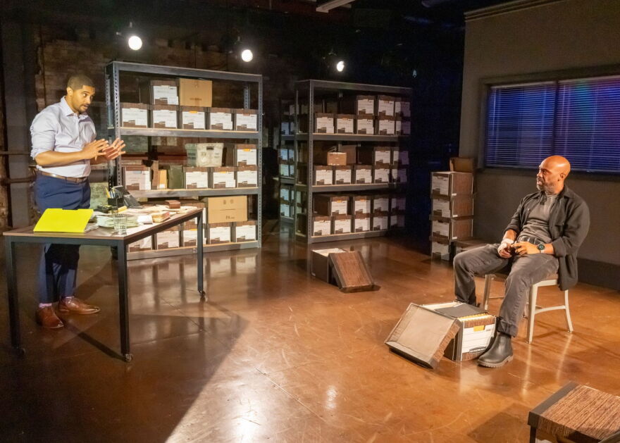 Two men stand on a stage, one behind a table and one in a chair across from him, with shelves filled with bankers' boxes in the background.