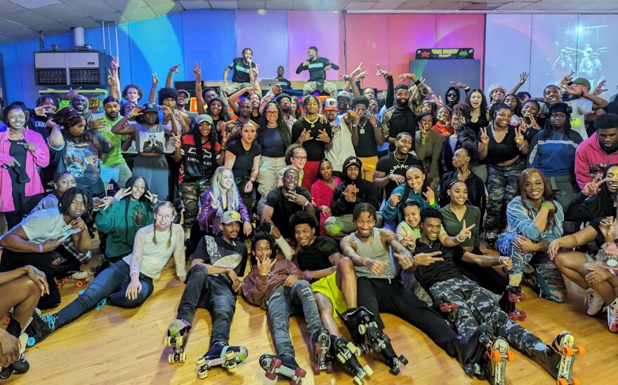 A large group of people stand, sit and lie on a floor inside a skating rink posing for the camera. 