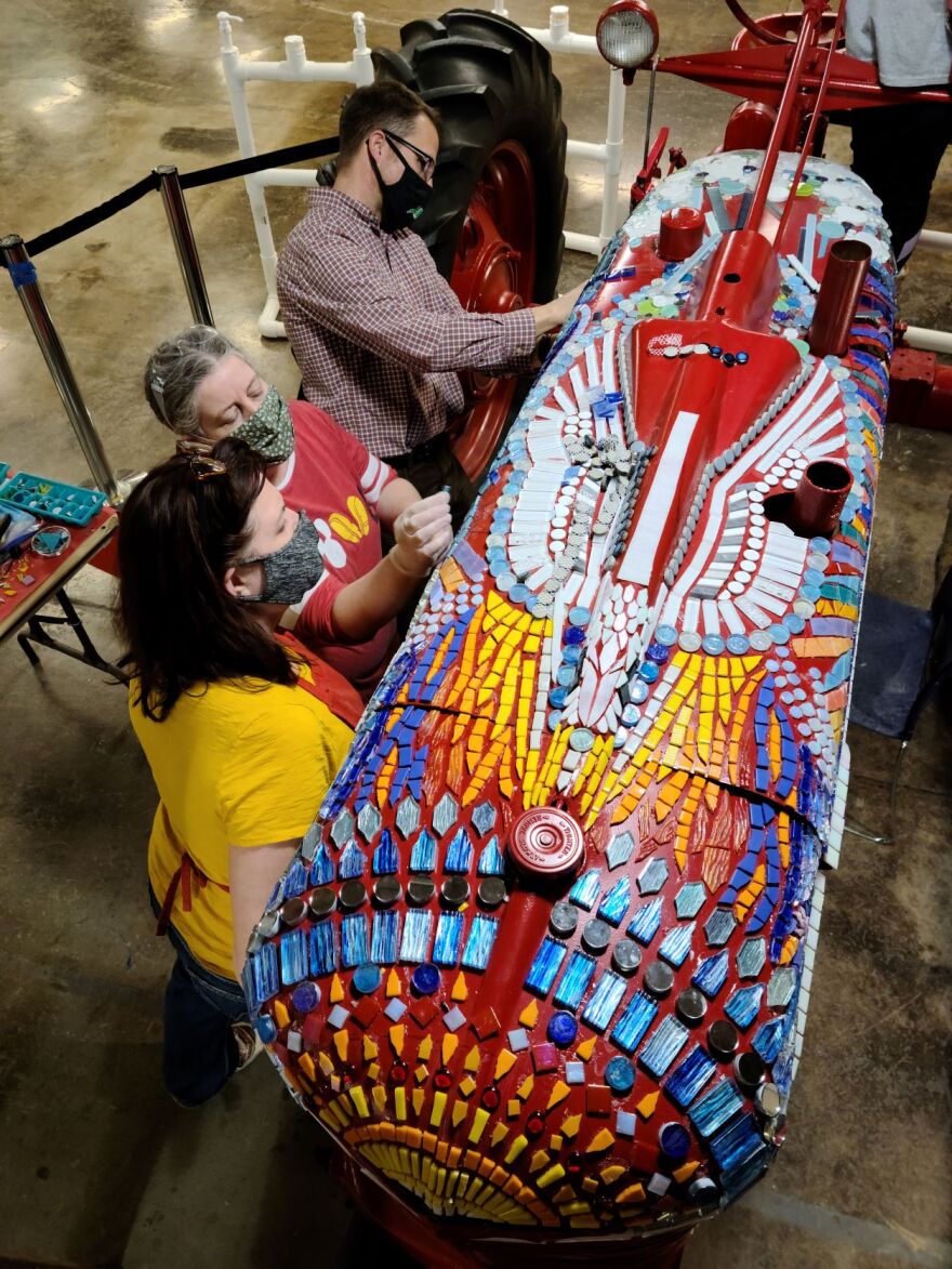 Three people glue small tiles on to a red tractor.