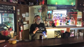 A woman wearing a mask mixes a drink wbehind a bar with a TV playing on the background.