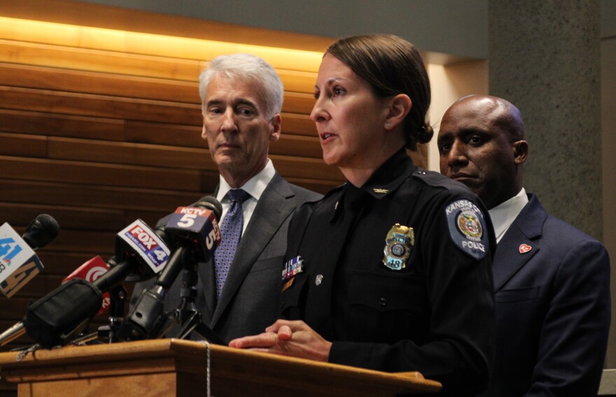 A woman in a police uniform stands at a podium. Two men stand behind her. 