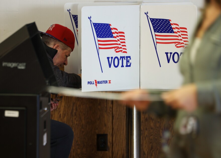 A man sits at a voting booth shielded by white dividers with the American flag and the word, "Vote."