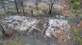 Charred ruins in grey ash sit on wet ground surrounded by trees.