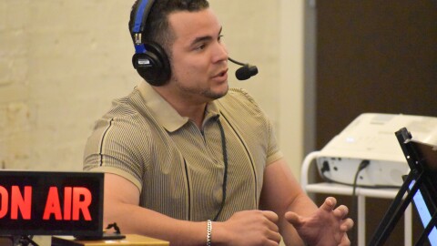 A man sits at a table with an "on-air" sign in front of him. The man is wearing a headphone and microphone set on his head. 