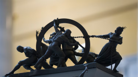 A close up of a mini model of people on top of a platform that reads "In God We Trust." The figures on top of the platform are pulling and pushing a hoop.